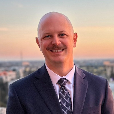 Timothy Gaull in a navy suit, smiling, with a city skyline at sunset behind him